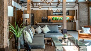 Un interior rústico con vigas de madera a la vista, una zona de estar central con cojines, una mesa de comedor y una cocina al fondo.
