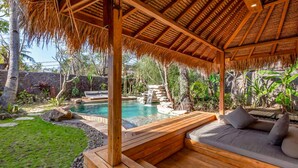 Una terraza de madera con techo de paja, una piscina con cascada y una zona de estar con cojines.