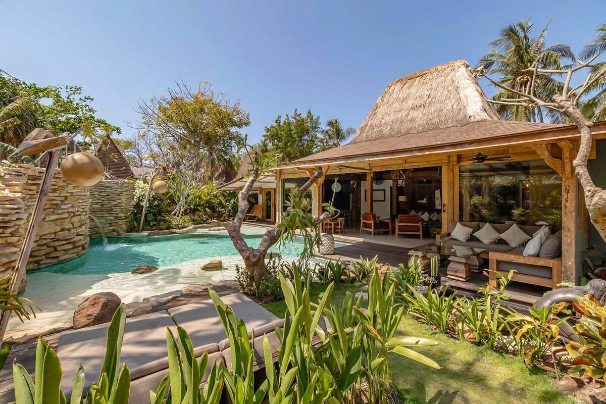 A poolside area with a thatched-roof structure, lounge seating, and a stone wall.