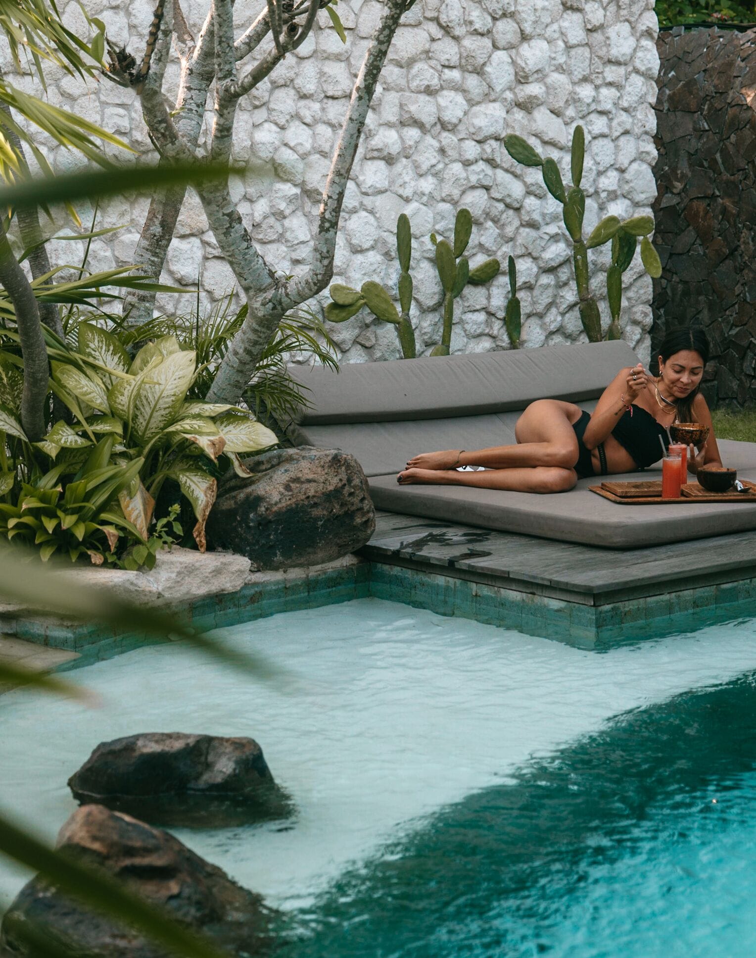Une personne se détend au bord d’une piscine, avec un mur de pierre et des plantes tropicales en arrière-plan.