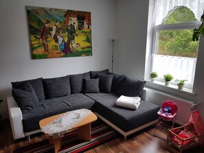 A living room with a dark grey sectional sofa, a small wooden table with a lace tablecloth, a window with potted plants, and a painting on the wall.