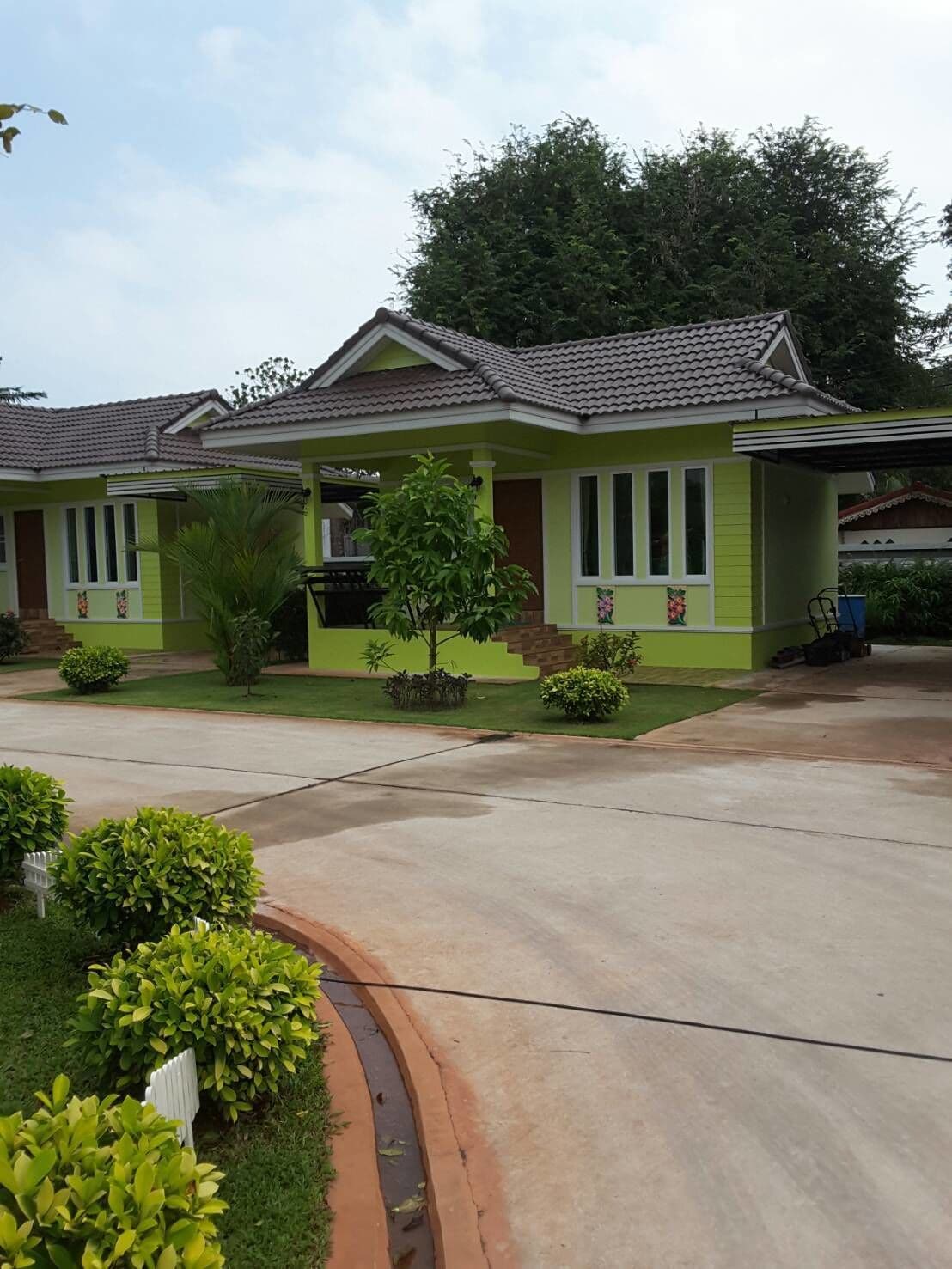 Uma casa de dois andares com telhado de telhas, paredes externas verdes e varanda frontal com uma pequena árvore e plantas em vasos.