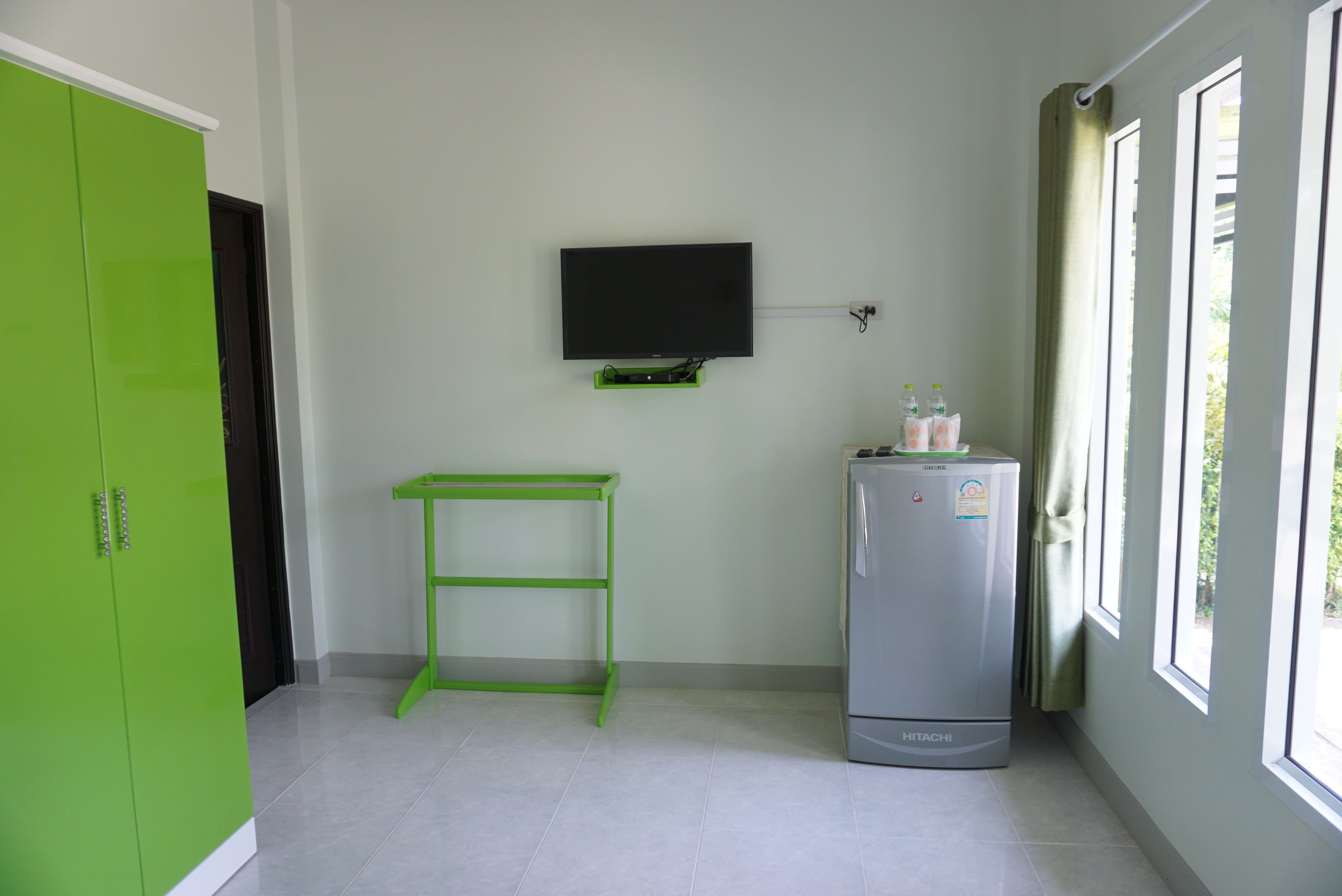 A room with a green wardrobe, a wall-mounted TV, a Hitachi refrigerator, and a window with curtains.