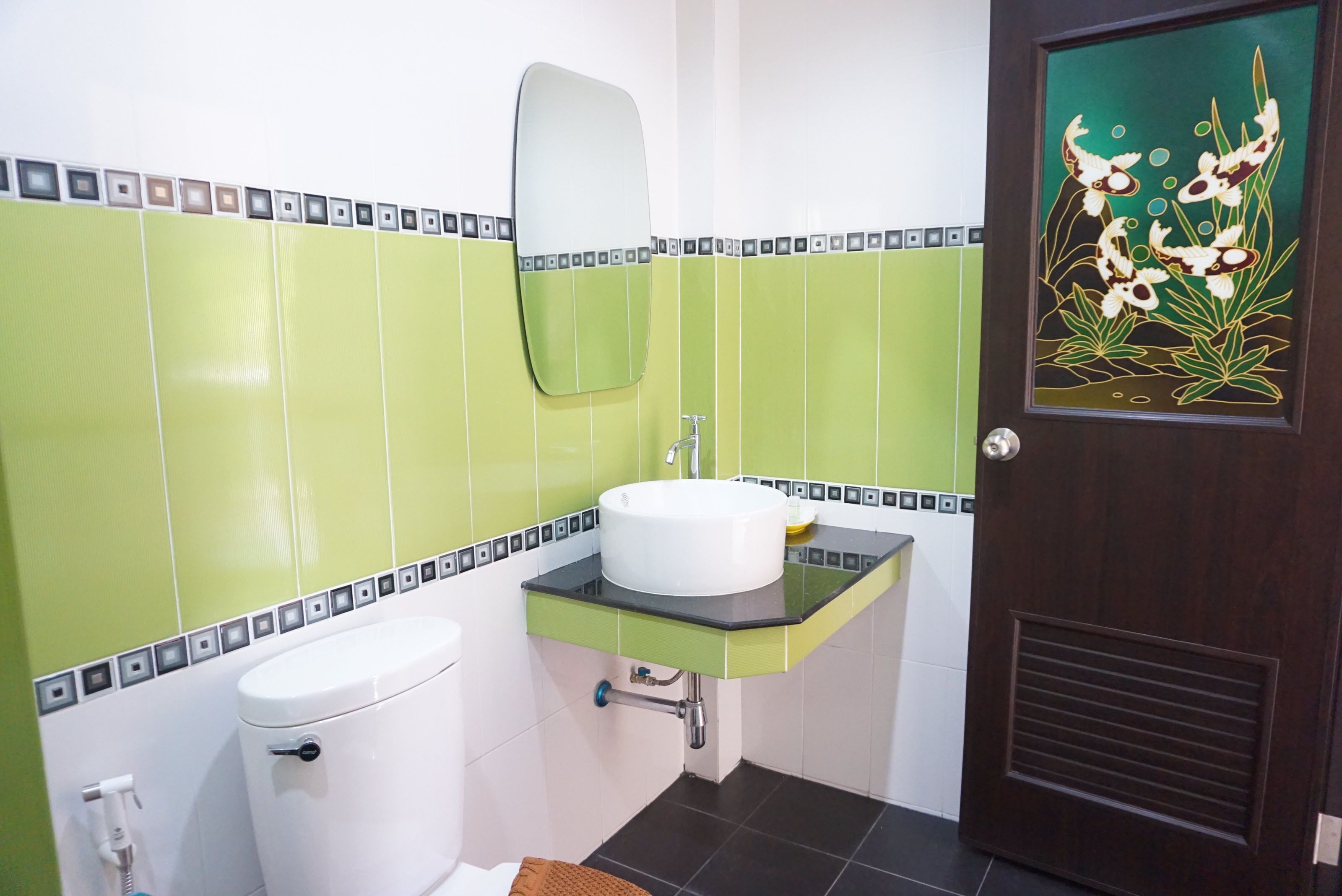 A bathroom with green and white tiles, a white toilet, a sink with a black countertop, and a mirror above the sink.