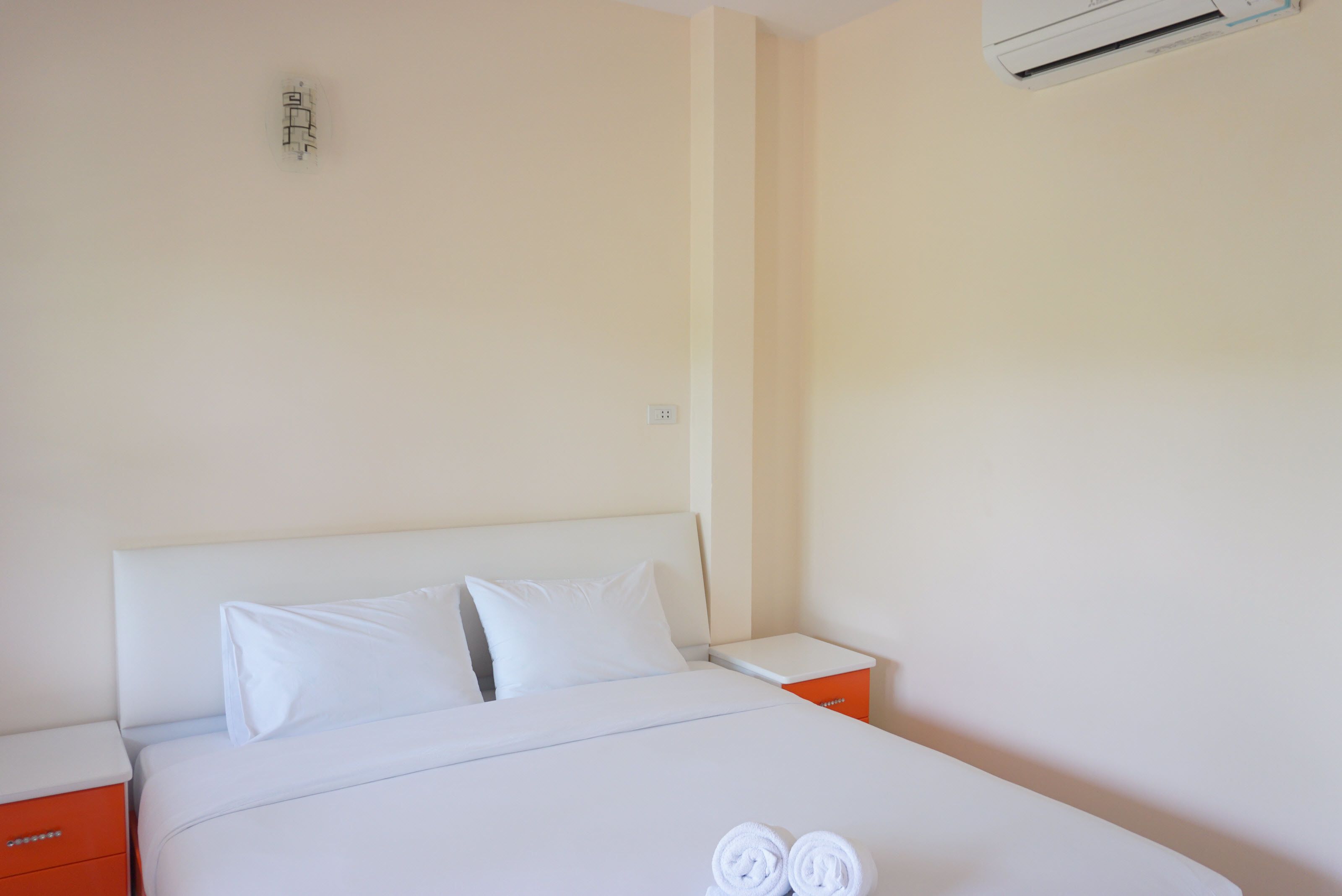 A hotel room with a bed, white linens, two pillows, a nightstand, an orange drawer, and an air conditioning unit.