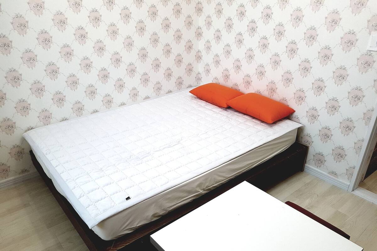 A bed with a white quilt and orange pillows, a small table, and patterned wallpaper.