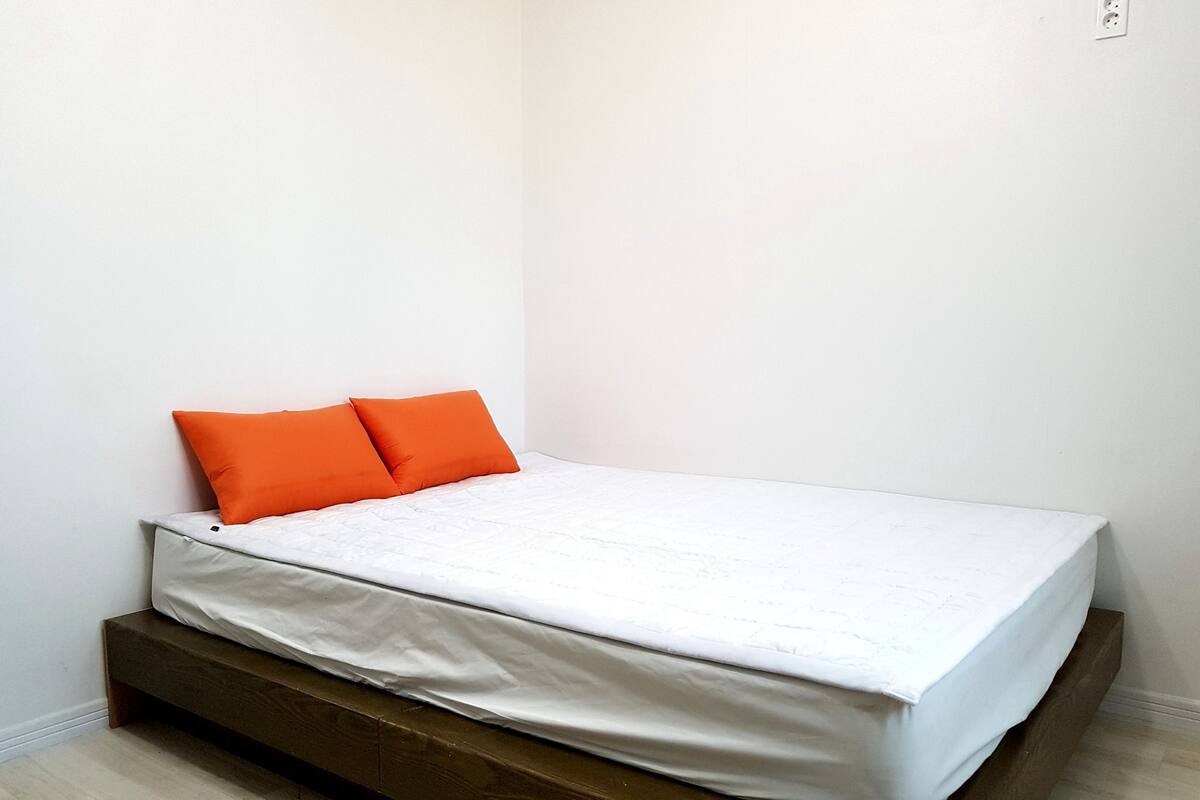 A small, single-bed room with a wooden platform bed, white bedding, and two orange pillows.