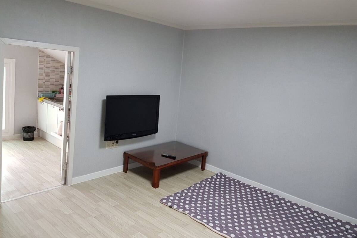 A room with a flat-screen TV mounted on the wall, a small wooden table, a patterned rug, and a light-colored wooden floor.