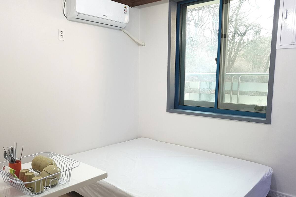 A small, single-bed room with a window, a white bedside table with a dish rack, and an air conditioning unit mounted on the wall.