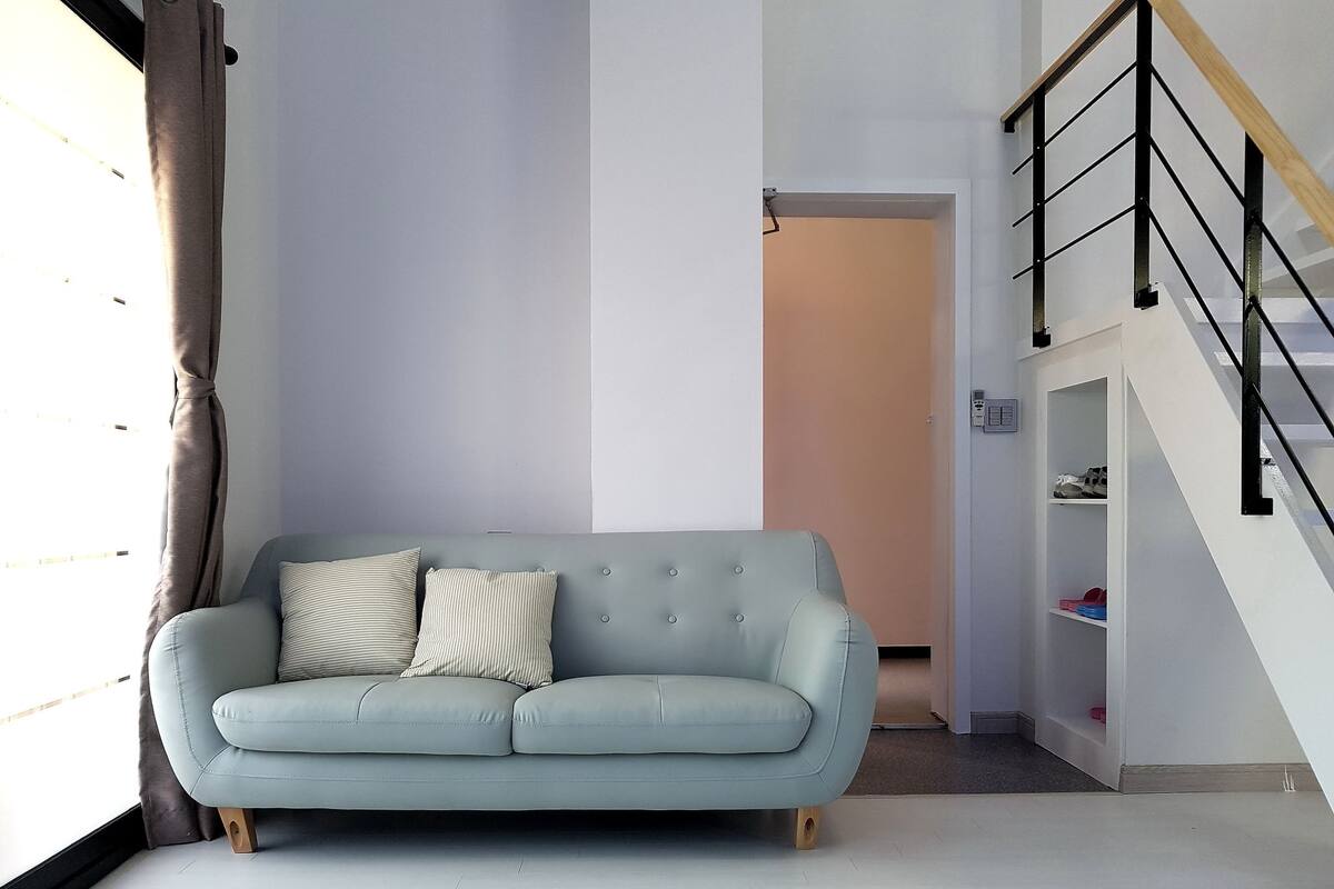 A light blue sofa with two pillows, a wooden floor, a staircase with black railings, and a white wall.