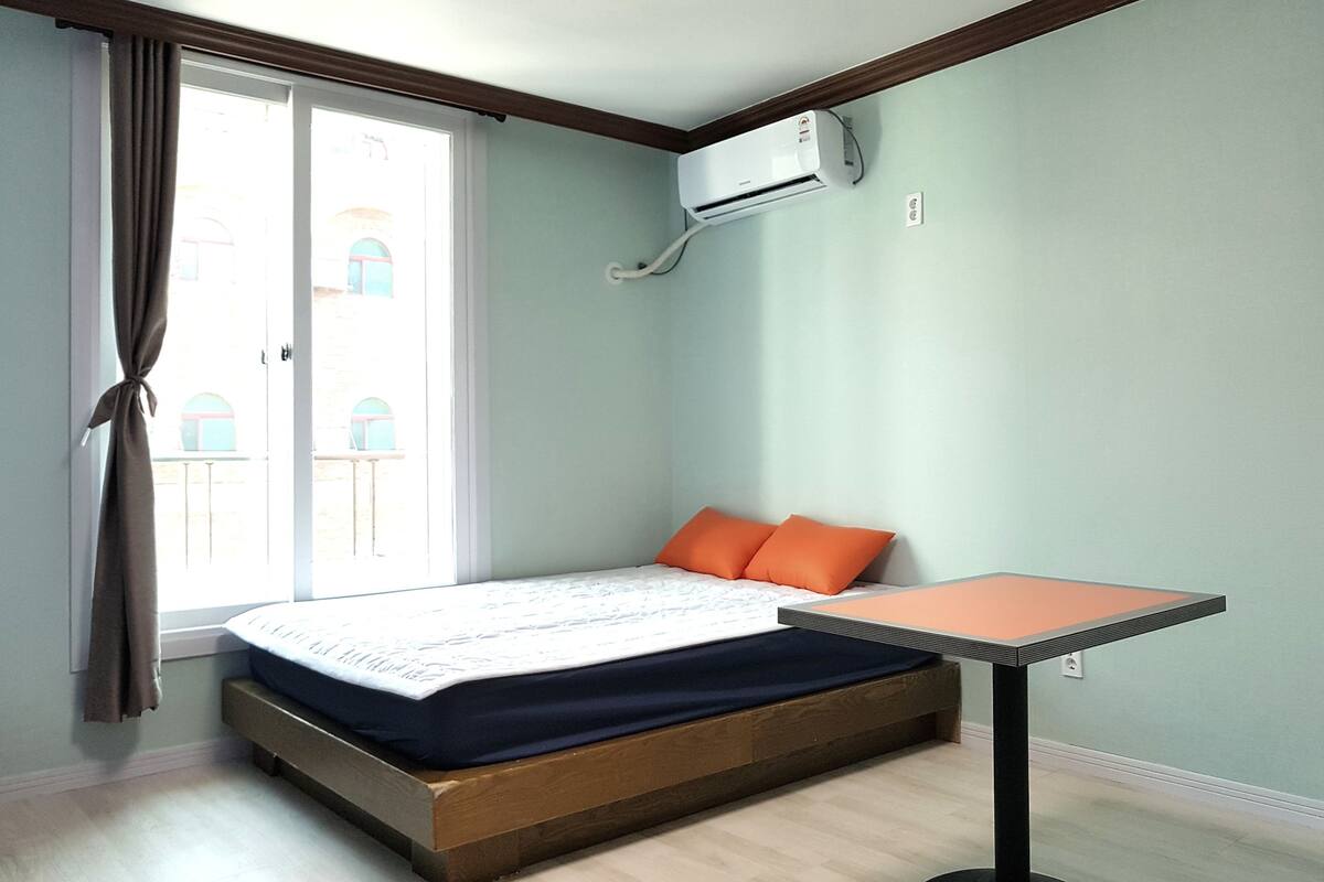 A bedroom with a bed, a table, an air conditioner, and a window with curtains.