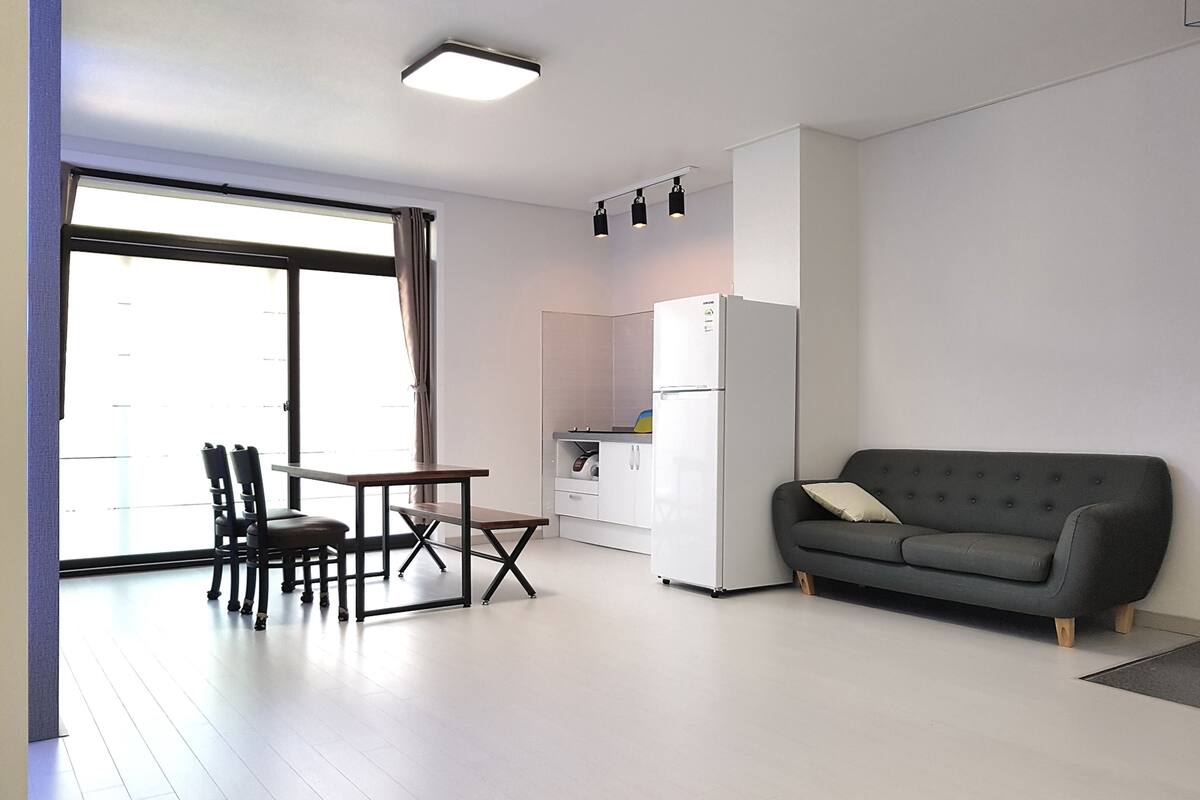 A modern living room with a grey sofa, a dining table with chairs, a kitchen area with a refrigerator, and a sliding glass door leading to a balcony.