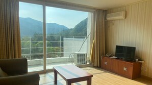 Een kamer met een grote schuifdeur naar een balkon met uitzicht op de bergen. Er staat een houten tafel, een bank en een televisie op een houten kast.