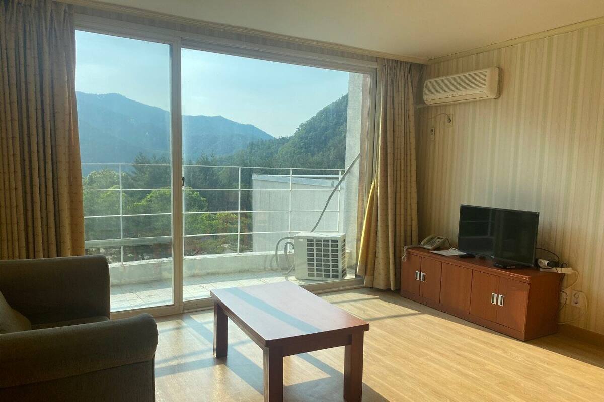 A room with a large sliding glass door leading to a balcony with a mountain view. There is a wooden table, a sofa, and a TV on a wooden cabinet.