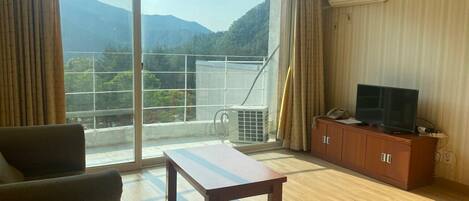 A room with a large sliding glass door leading to a balcony with a mountain view. There is a wooden table, a sofa, and a TV on a wooden cabinet.
