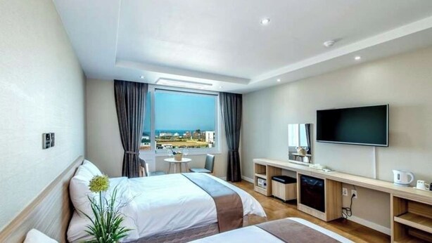 A hotel room with two beds, a TV, a desk with a chair, a small table, and a view of the city. - Jeju IN Hotel (Jeju City)