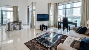 A modern living room with a glass coffee table, a sofa, a TV, and a view of a cityscape and water. - Park Island - Bonaire by Deluxe Holiday Homes (Dubai)