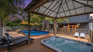 A wooden deck with a hot tub and a swimming pool, surrounded by lounge chairs and a covered area with a railing. - Elk Estate (Inverloch)