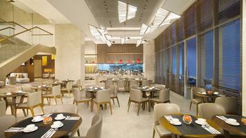 A modern hotel dining area with multiple tables set for dining, a staircase, and a view of the kitchen in the background.
