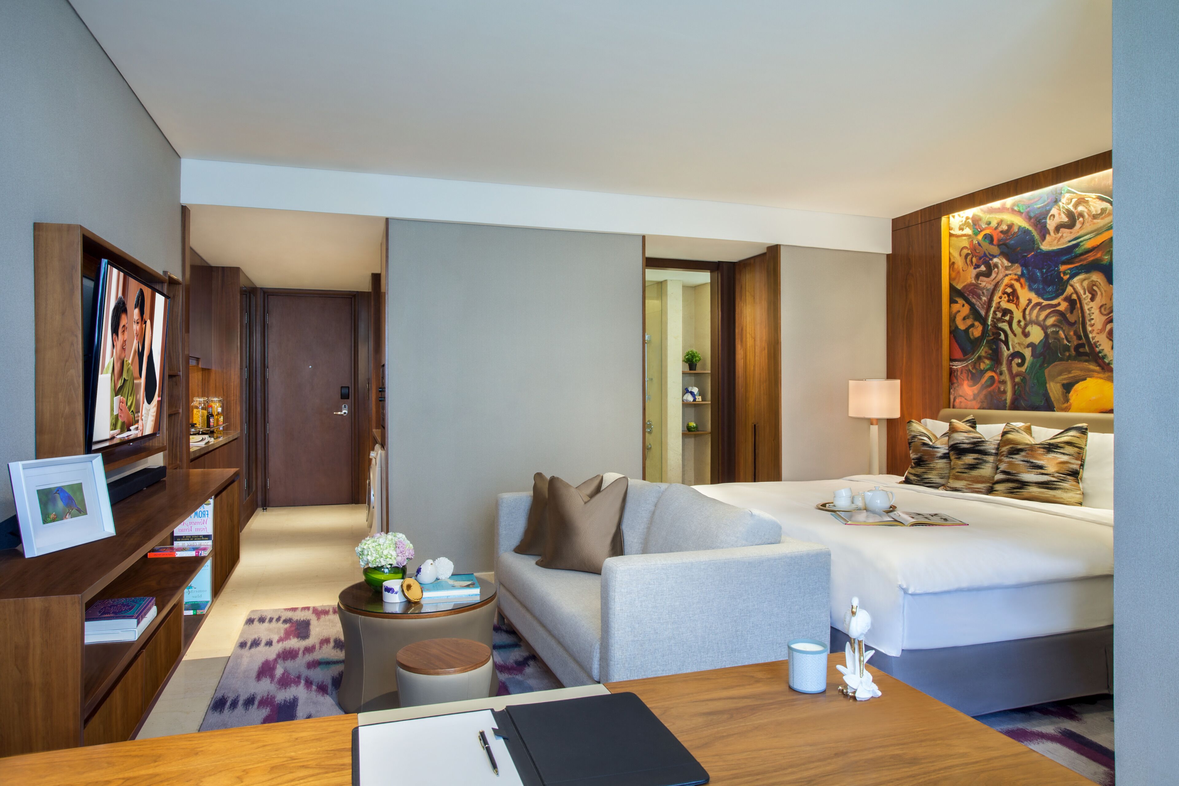 Una habitación de hotel con sofá, cama, escritorio, estantería y un cuadro en la pared.