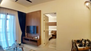A modern hotel room with a flat-screen TV mounted on a wooden panel, a small table with a vase of flowers, and a dining area with a stove. - Roomy at Bukit Bintang (Kuala Lumpur)