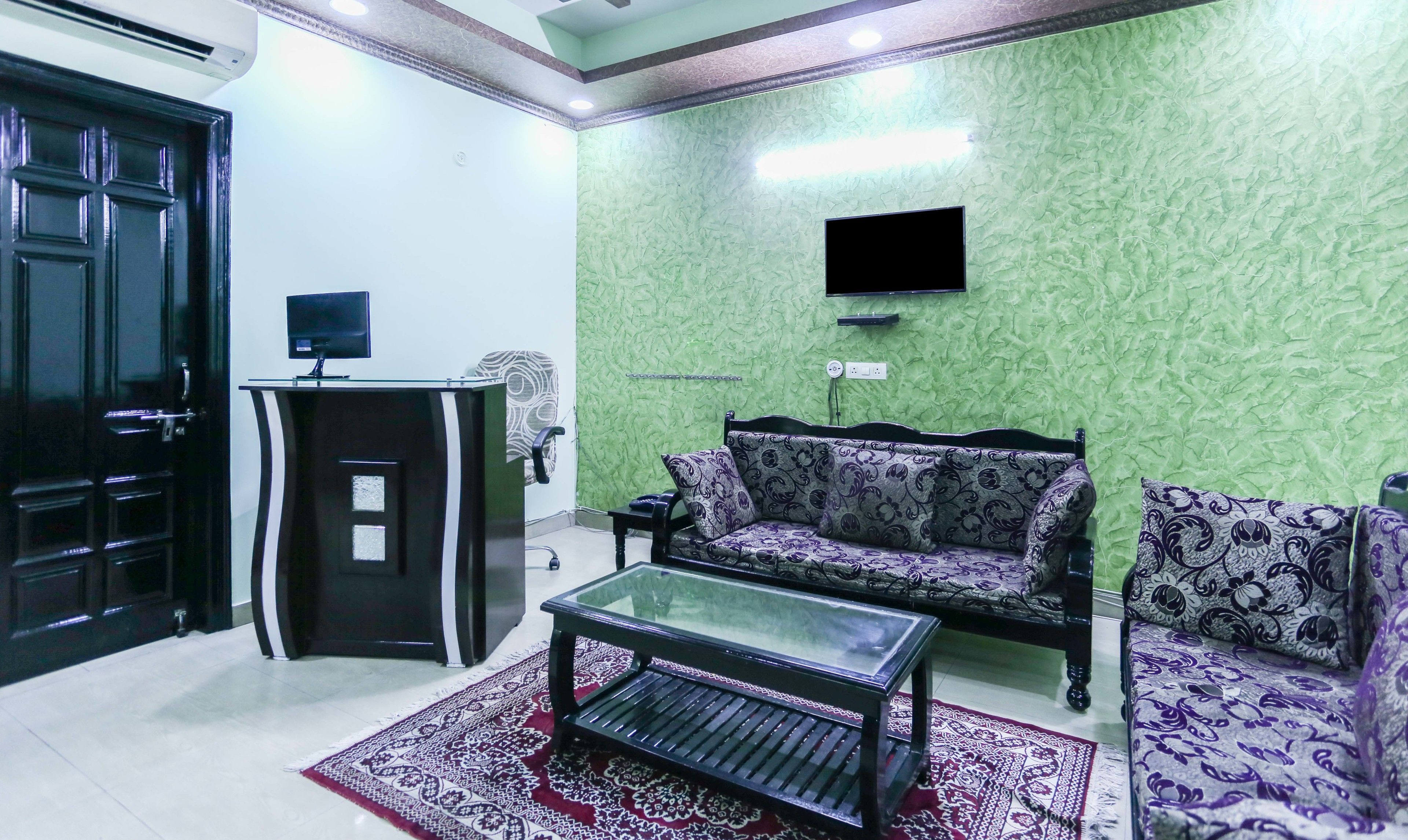 A living room with a green textured wall, a black sofa, a patterned armchair, a glass coffee table, and a black cabinet with a flat-screen TV mounted above it.
