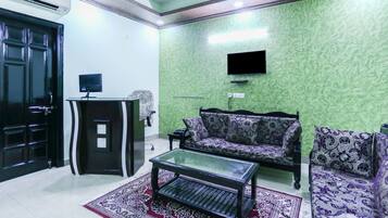 A living room with a green textured wall, a black sofa, a patterned armchair, a glass coffee table, and a black cabinet with a flat-screen TV mounted above it.