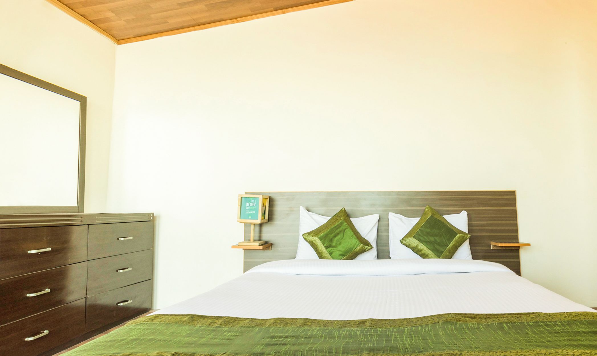 A neatly made bed with a green bedspread and two pillows, a wooden nightstand with a clock, and a dark wooden dresser with multiple drawers.