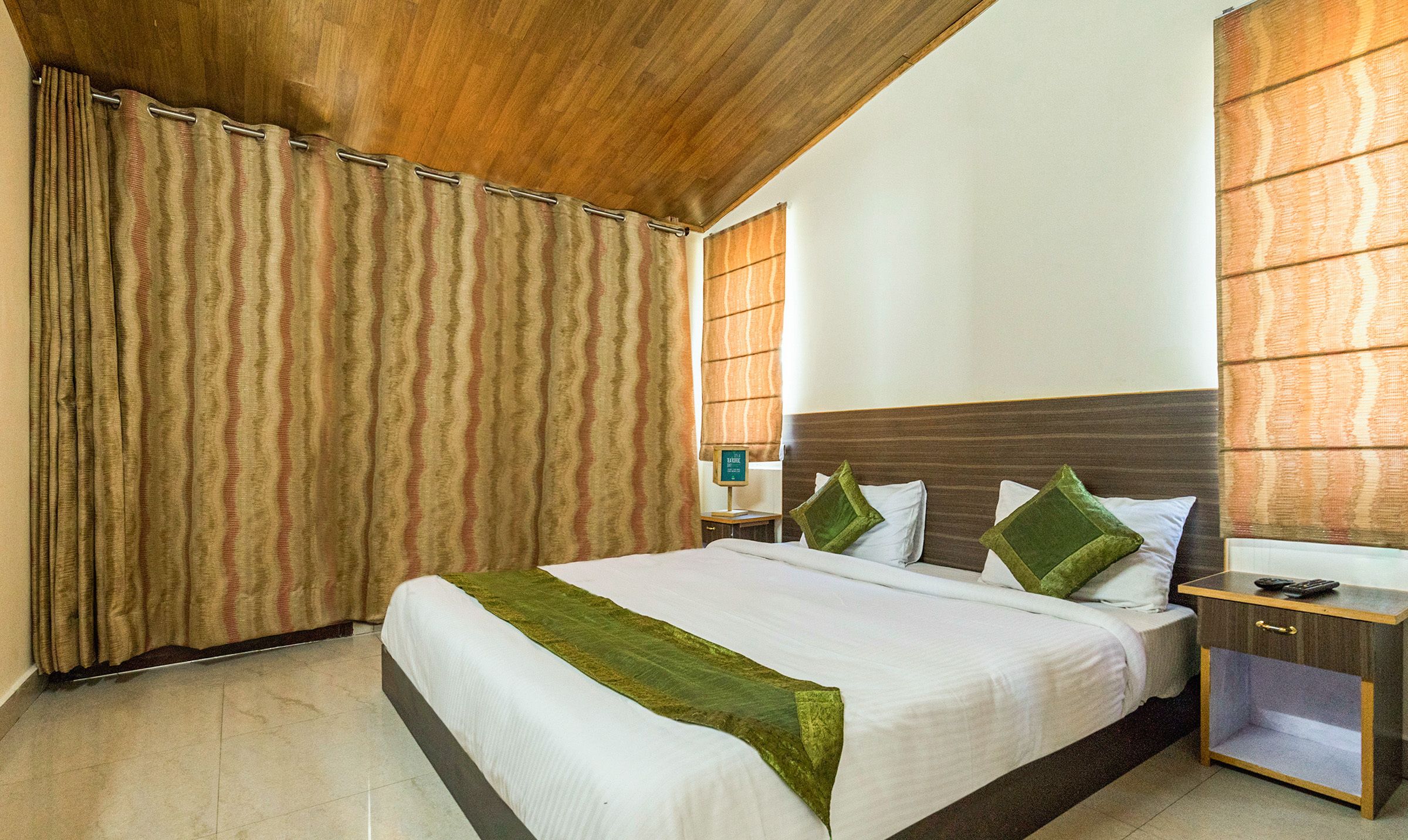 Quarto de hotel com cama grande, mesa de cabeceira, cabeceira de madeira e janela com persianas.