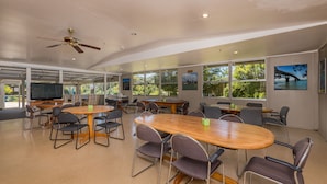 A spacious conference room with multiple tables and chairs, large windows with a view of greenery, and a ceiling fan. - Riverside Lodge Paihia (Haruru)