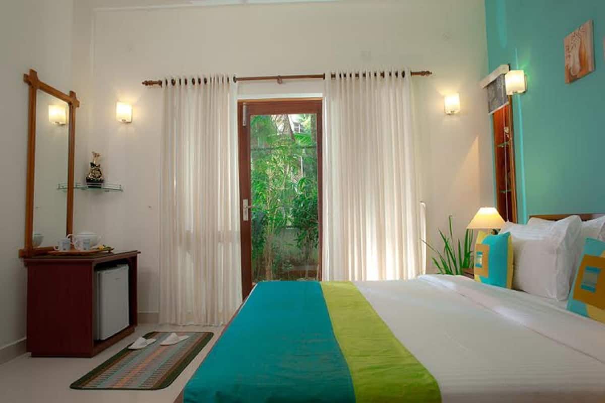 A hotel room with a bed, a small desk, a mirror, and a view of greenery through the door.