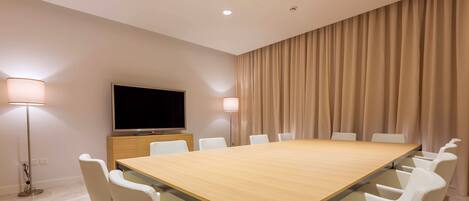 A conference room with a long table, white chairs, a flat-screen TV, and beige curtains.