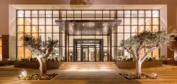 Una entrada de hotel moderna con puertas de cristal, un corredor cubierto y jardines simétricos.