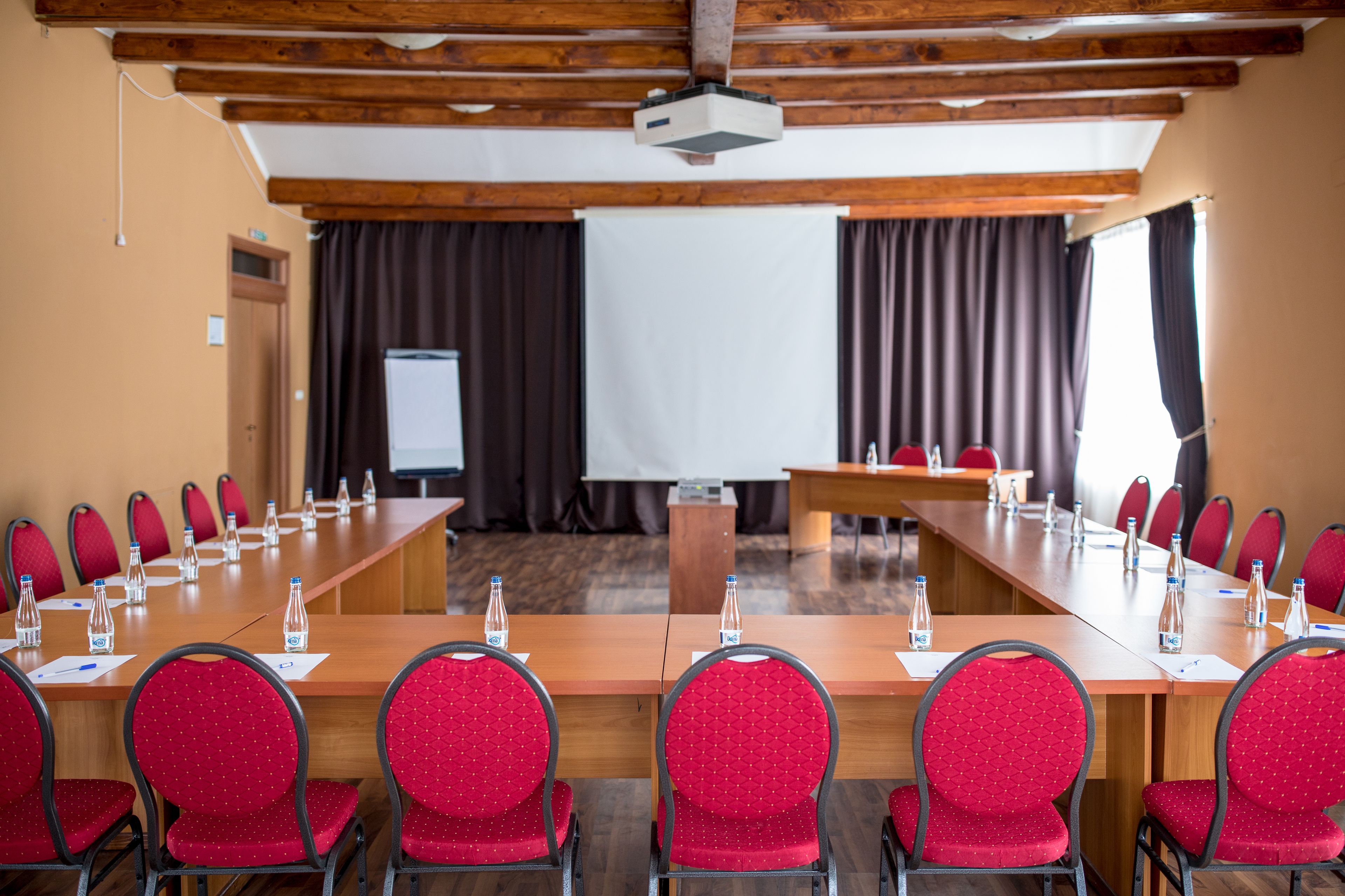A conference room with a long table, red chairs, water bottles, and a projector screen.