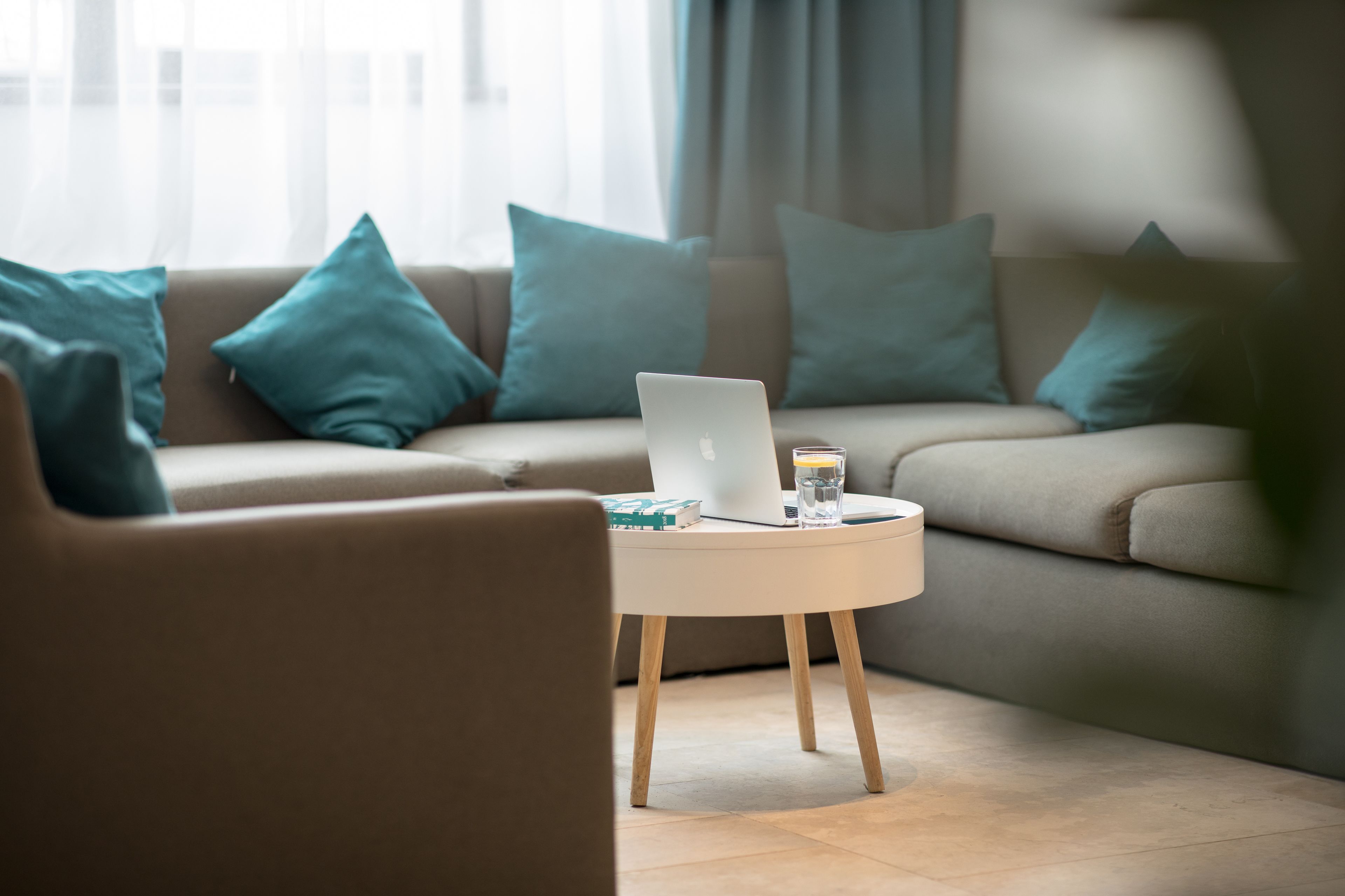 Una sala de estar moderna con un sofá color beige, cojines turquesa, una mesa redonda blanca con una computadora portátil, libros y un vaso de agua.