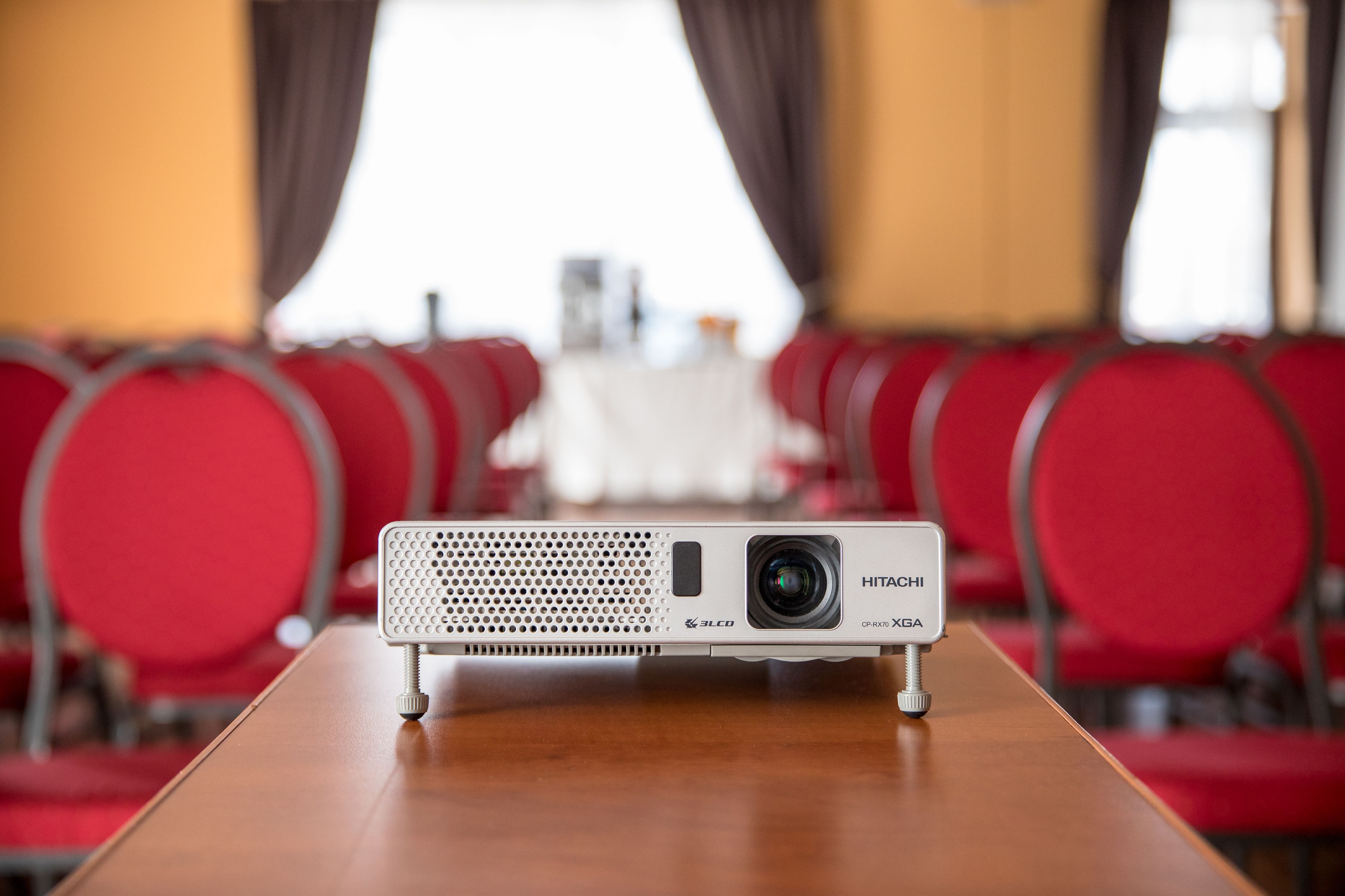 Un proyector Hitachi sobre una mesa de madera en una sala de conferencias con sillas rojas y una mesa blanca al fondo.