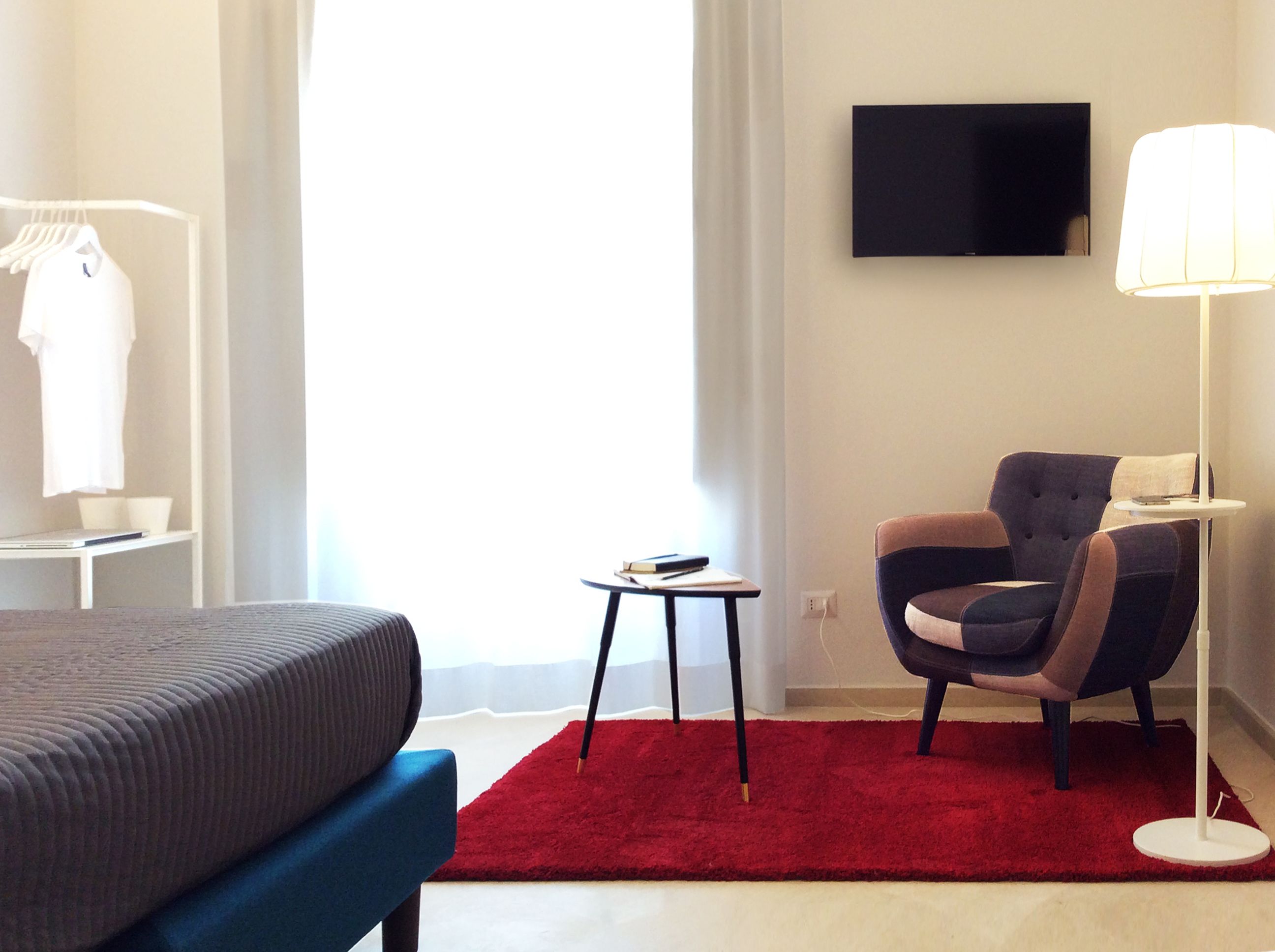 Habitación de hotel con cama, alfombra roja, una silla, una mesita, lámpara y televisor montado en la pared.