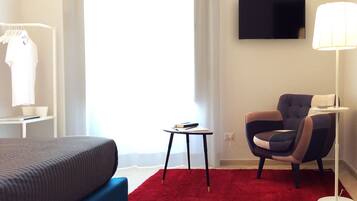 Habitación de hotel con cama, alfombra roja, una silla, una mesita, lámpara y televisor montado en la pared.