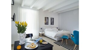 A modern hotel room with a bed, a blue armchair, a dining table with breakfast, and a vase of flowers. - Re Federico Boutique Hotel (Syracuse)