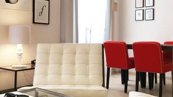 Una sala de estar moderna con un sofá capitonado, una mesa de centro de vidrio, una zona de comedor con sillas rojas y una araña.