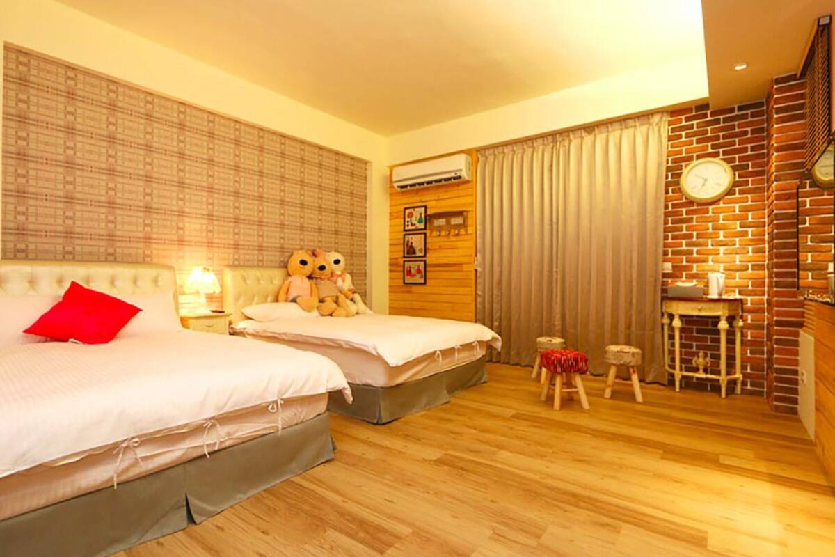 A hotel room with two beds, a small stool, a table with a clock, and a brick wall.