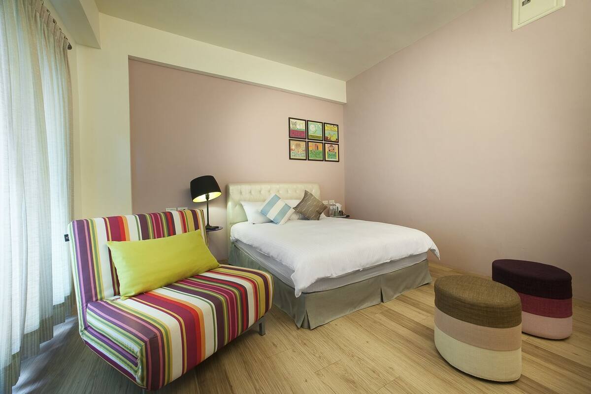 A hotel room with a bed, a colorful striped sofa, a bedside table with a lamp, and a small ottoman.