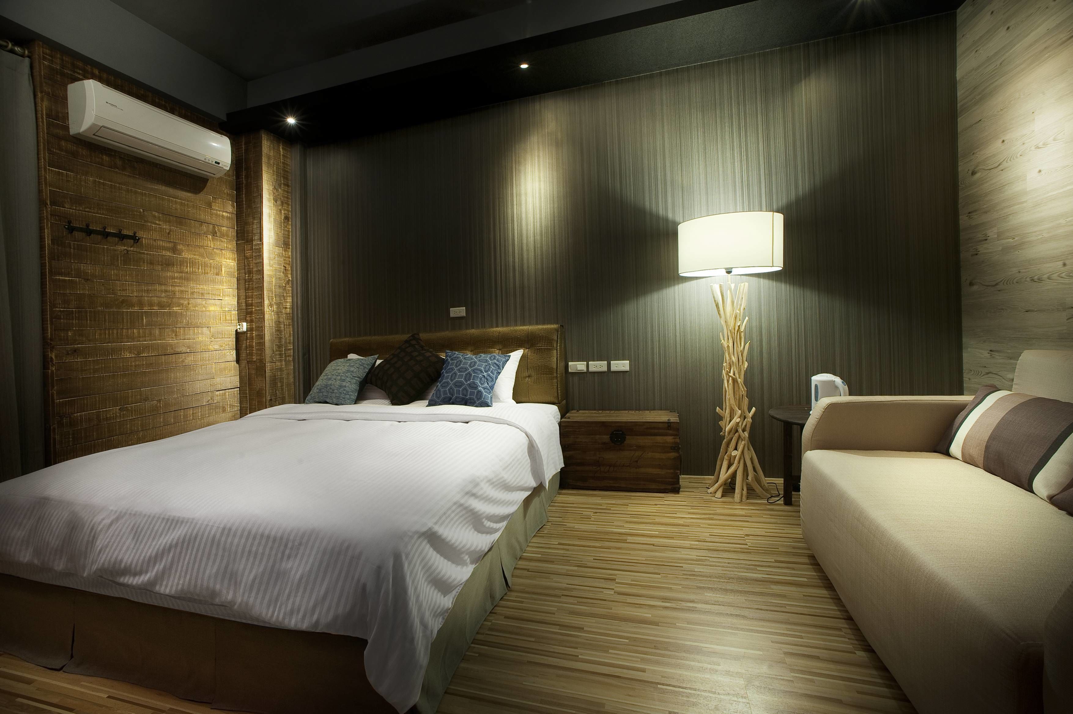 A modern hotel room with a large bed, a sofa, a bedside table, a lamp, and a wall-mounted air conditioner.