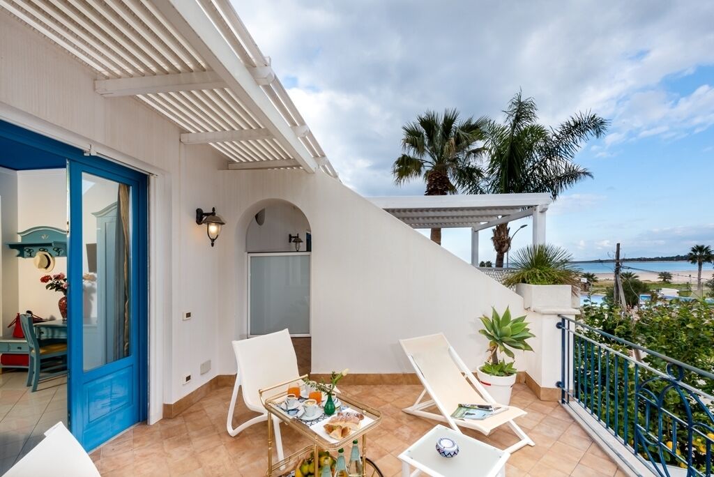 A balcony with two white lounge chairs, a small table with a tray of food and drinks, and a view of the beach.