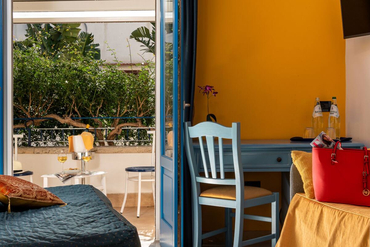 A room with a bed, a desk, a chair, a table, and a window overlooking greenery.