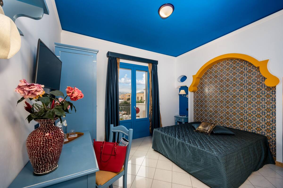 A bedroom with a blue ceiling, a bed with a patterned headboard, a desk with a vase of flowers, and a view through a door.