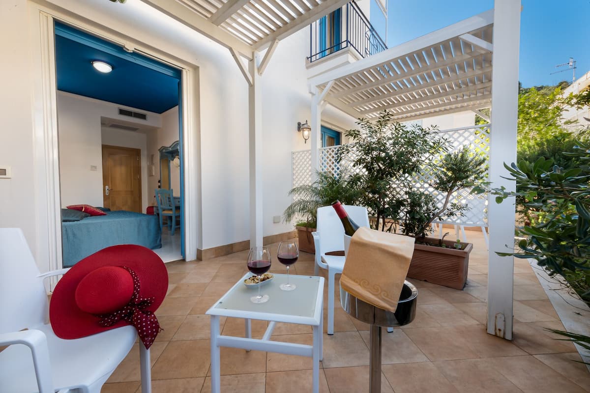 A patio area with a white table, chairs, and a red hat on a chair. There is a potted plant and a bottle of wine on the table.