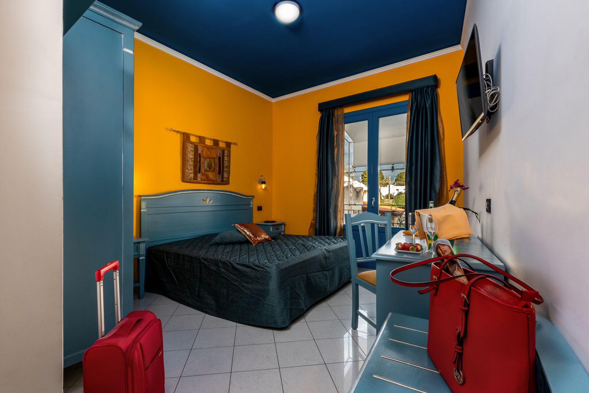 A hotel room with a bed, a desk with a chair, a suitcase, and a TV mounted on the wall.