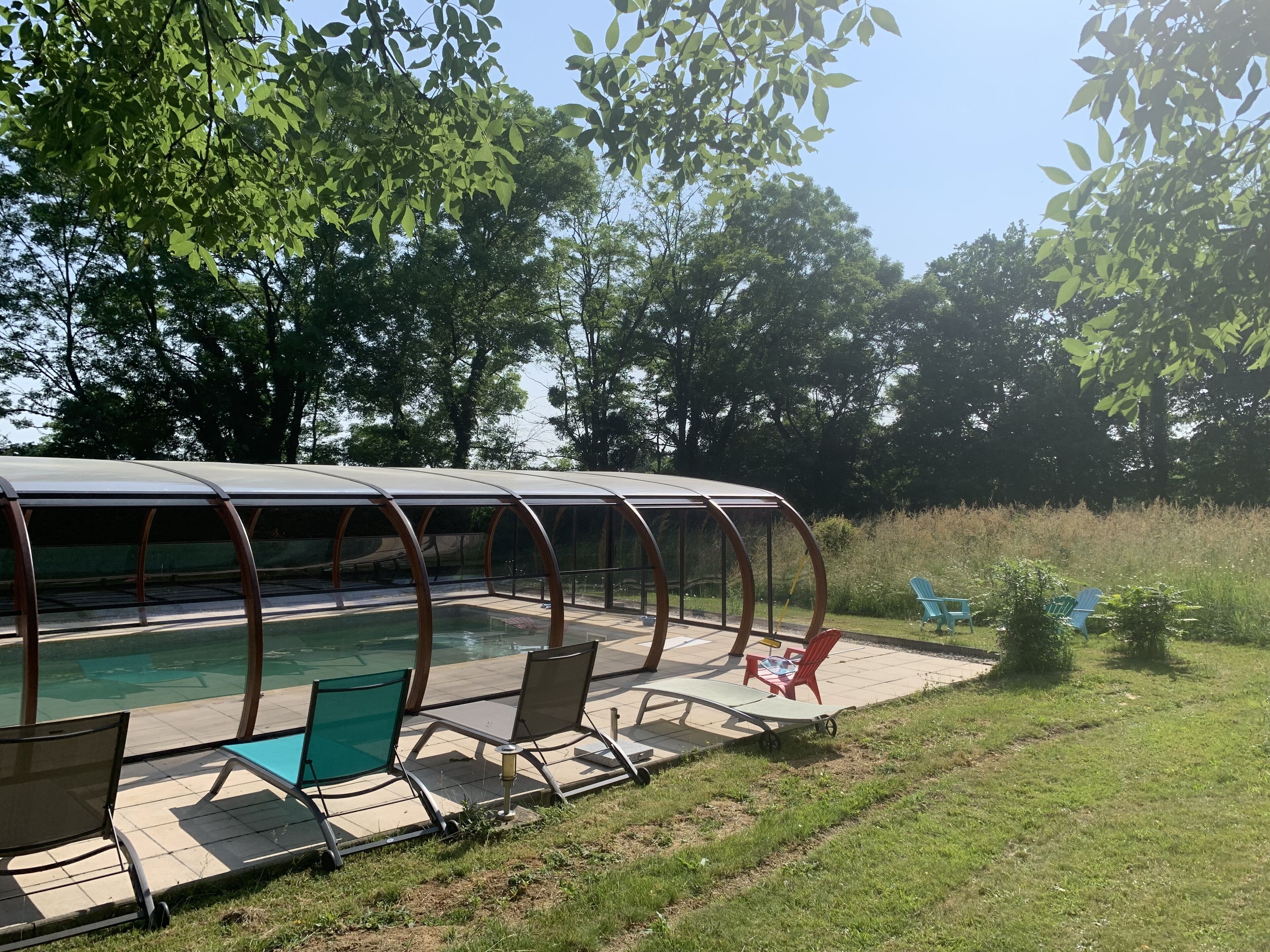 Une piscine extérieure couverte, dotée de chaises longues et d’une table, et entourée d’arbres et d’herbe.