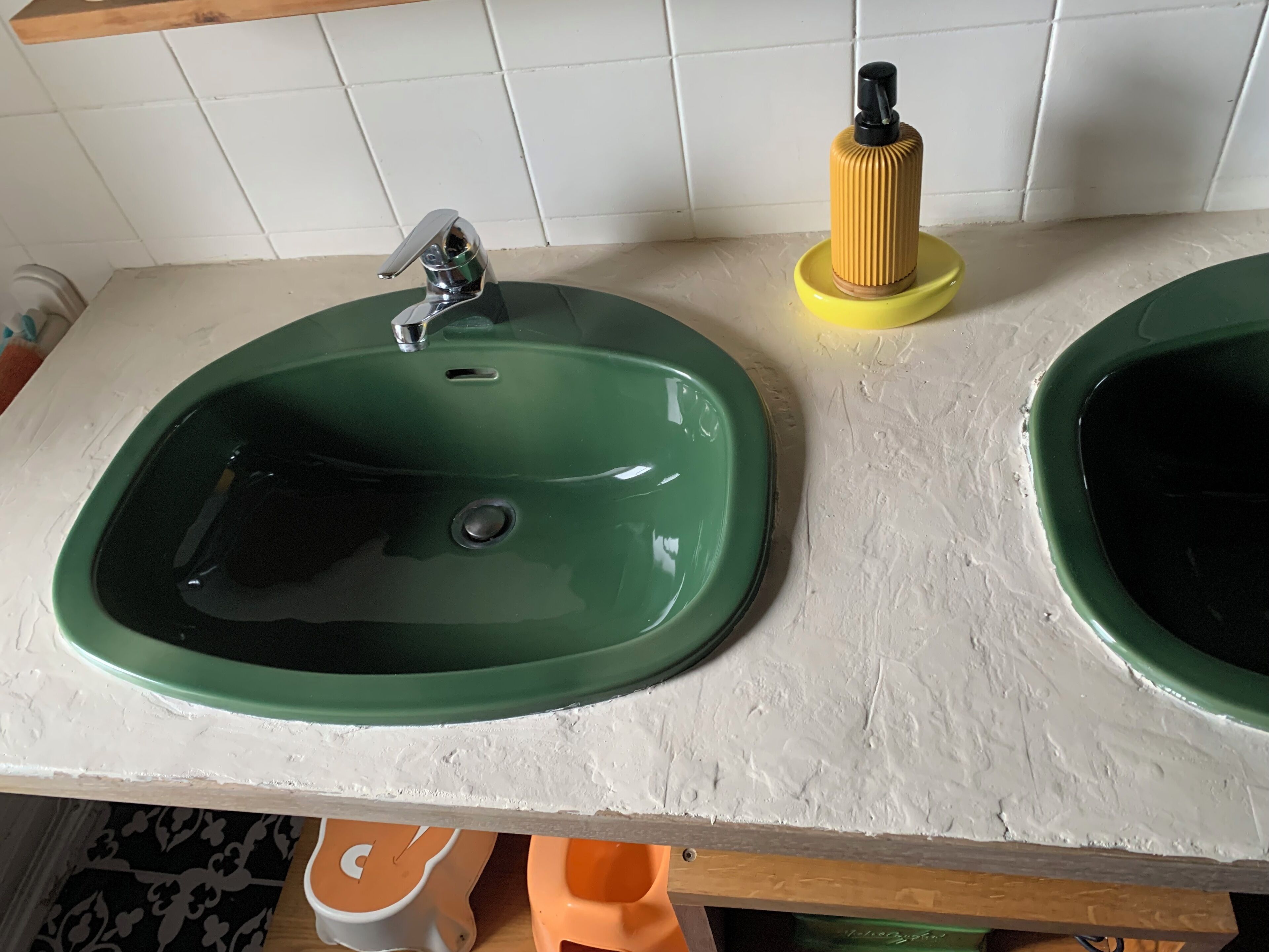 Un baño con dos lavamanos verdes, un dispensador de jabón amarillo y una pared de azulejos blancos.